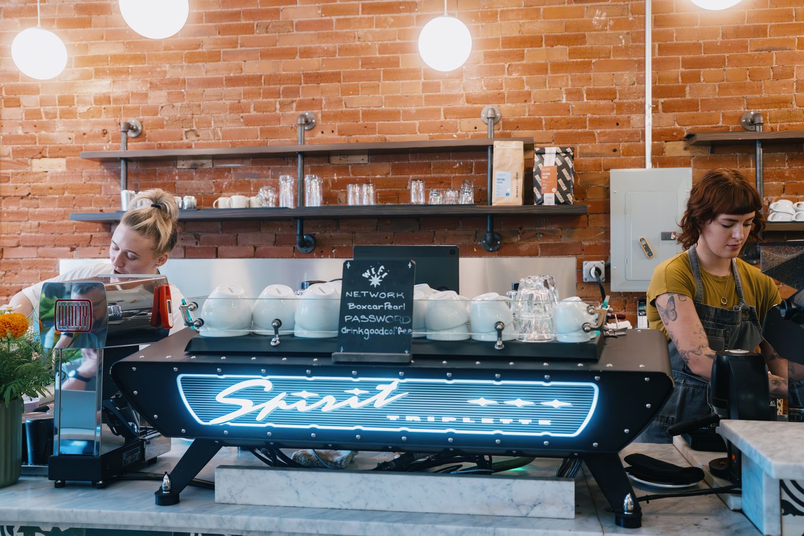 Two Boxcar baristas working behind a lit Spirit Triplette espresso machine in front of a brick wall