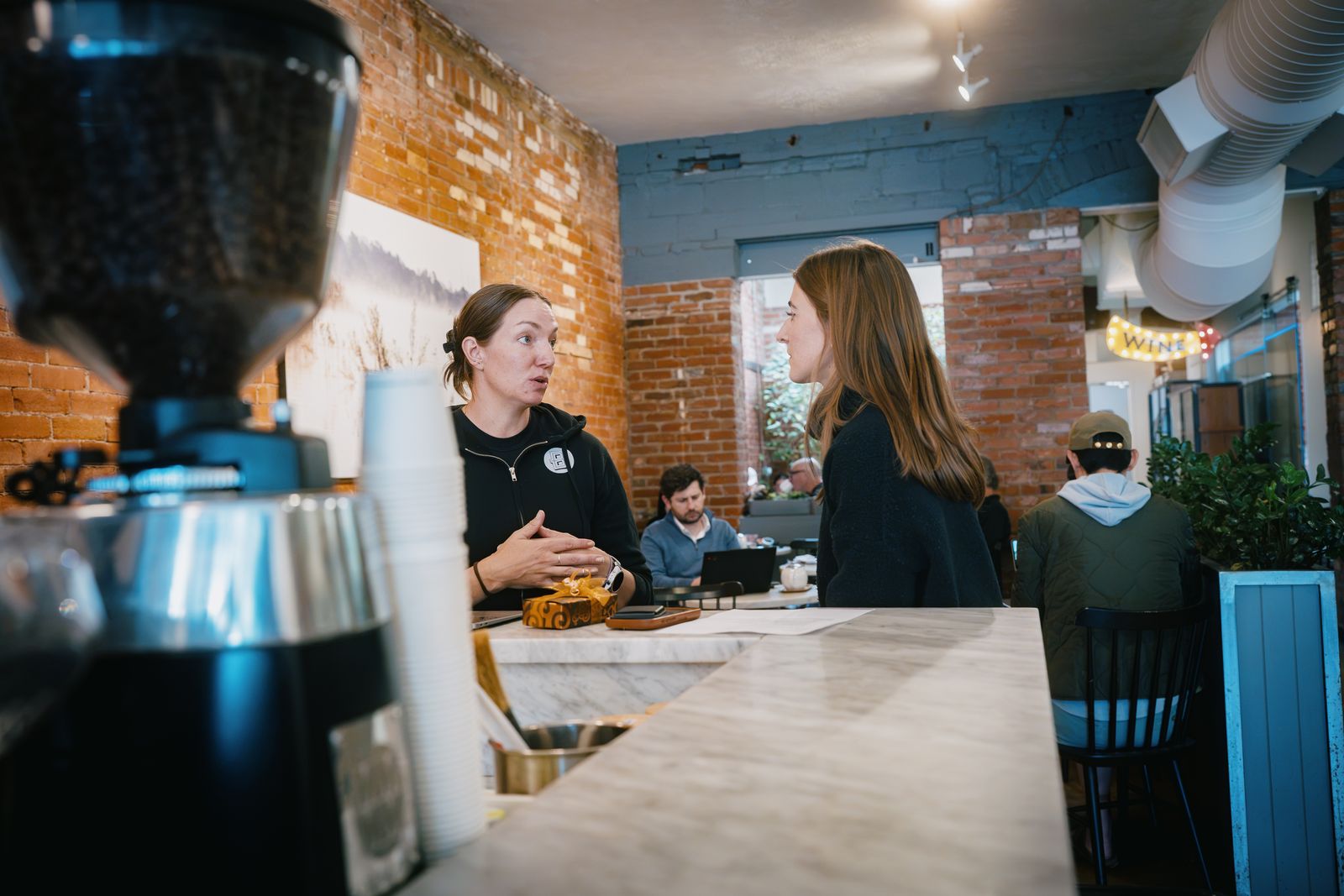 Linnea of Boxcar Coffee Roasters talking with Willow concierge Morgan Trego across the counter at Boxcar while customers work in the background