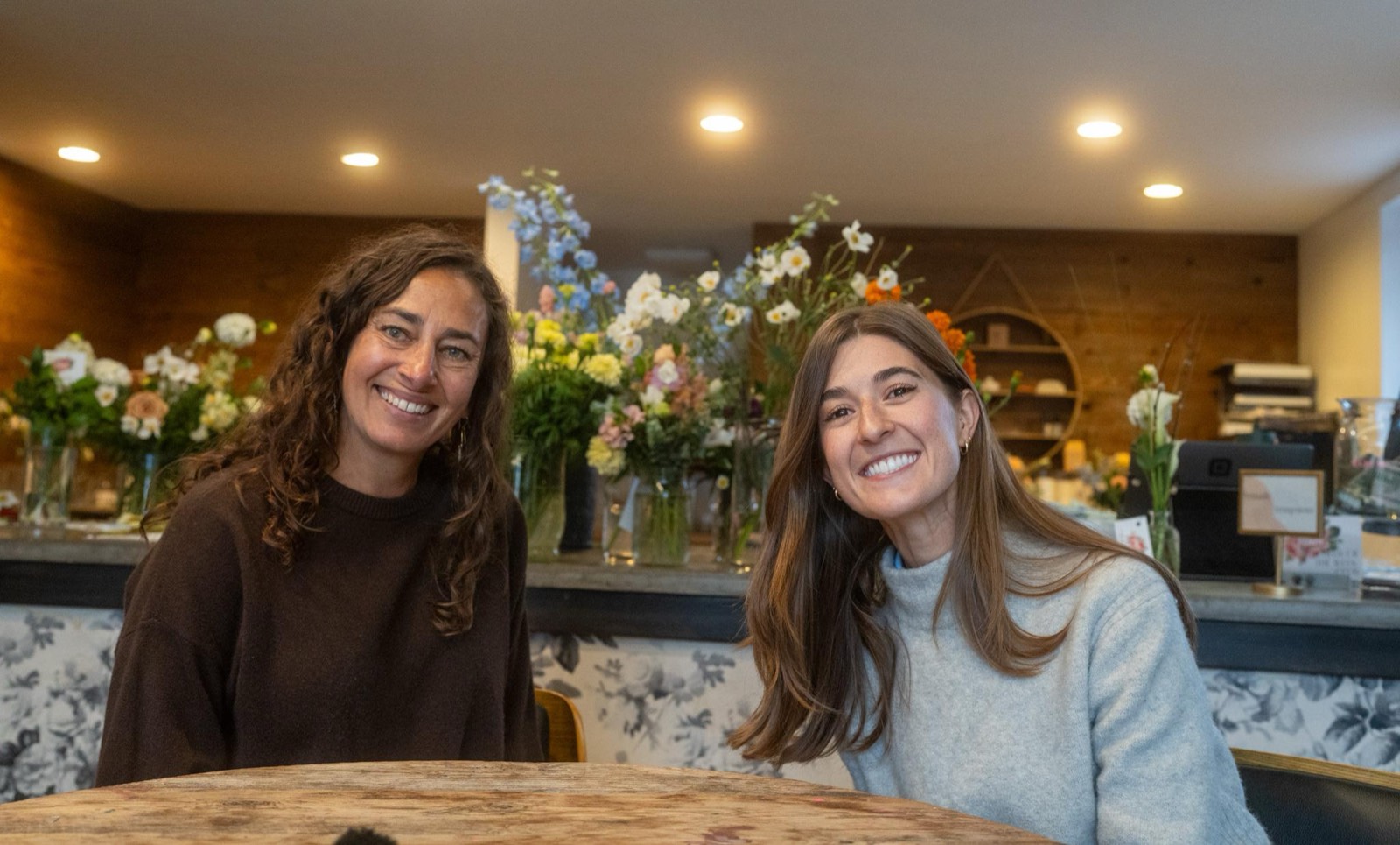 Lara of Fiori Flowers and Willow concierge Morgan Trego smiling at a table inside the shop, with fresh arrangements on the counter behind them